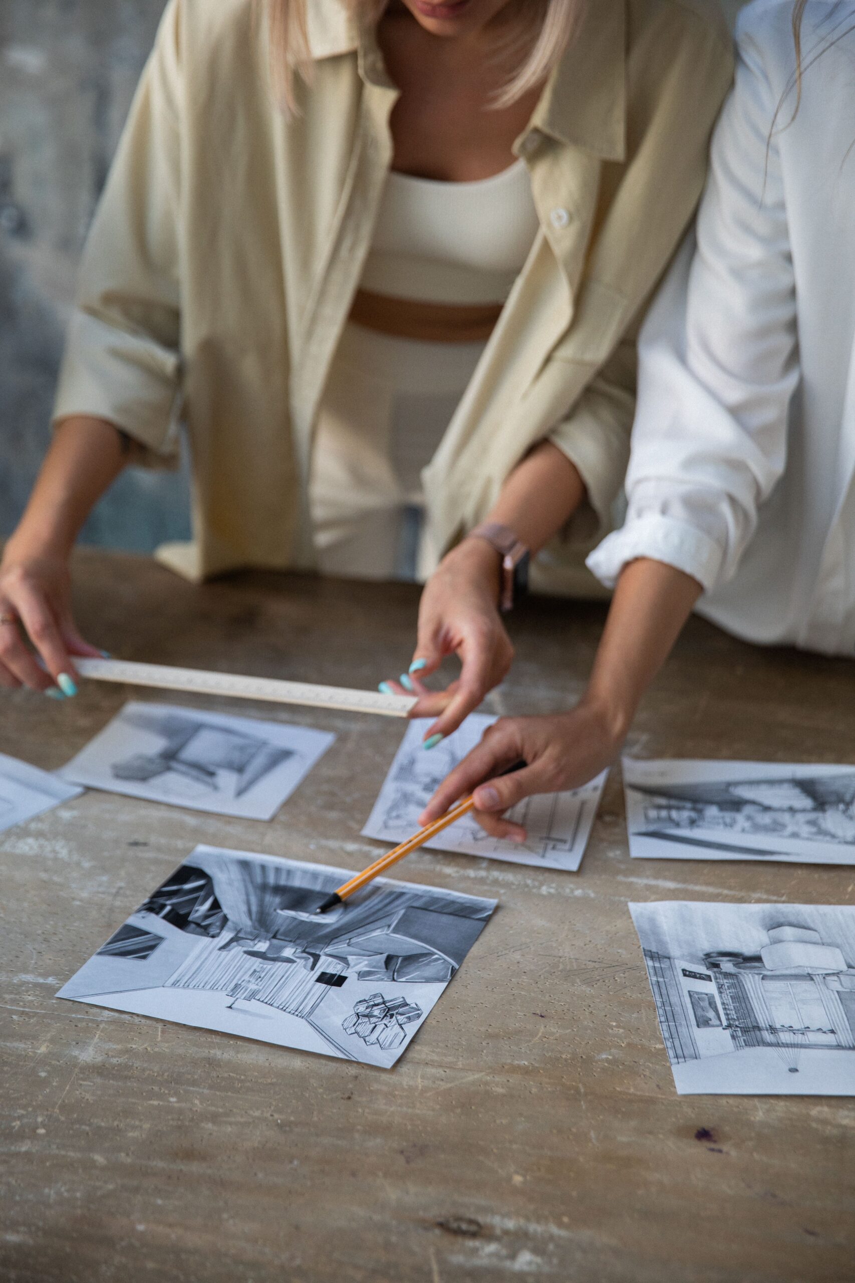 Two women collaborating in an art studio, examining design sketches on a wooden table.
